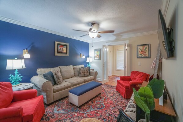 A living room with a blue wall, beige sofa, red chairs, a ceiling fan, and a patterned rug. - Stylish 2BR Condo w/ Pool, Fitness Center, Walk to Shops & Dining (St. Simons Island)