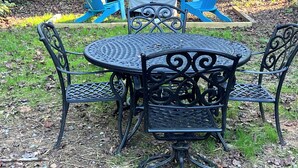 A round metal table with four chairs, surrounded by a brick wall and blue Adirondack chairs in the background.