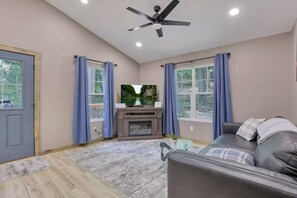 A living room with a grey leather sofa, a flat-screen TV, a fireplace, and large windows with blue curtains. - Tranquil Ridge Retreat | Hocking Hills (Logan)