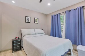 Un dormitorio con cama, mesita de noche, ventana con cortinas y dos cuadros en la pared.