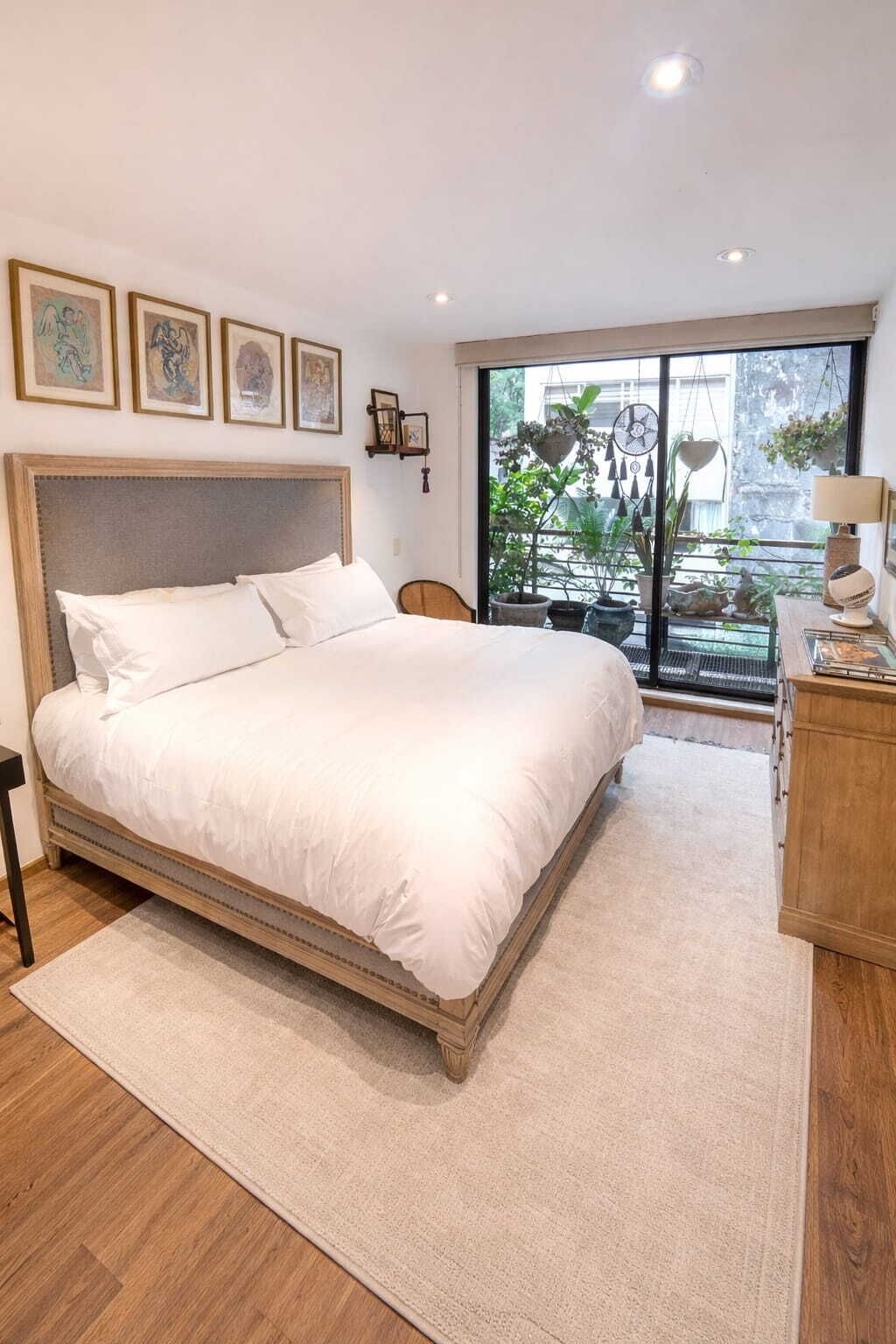 Une chambre à coucher avec un grand lit, une tête de lit en bois, une table de chevet, une chaise, une fenêtre donnant sur des plantes et des tableaux encadrés accrochés au mur.