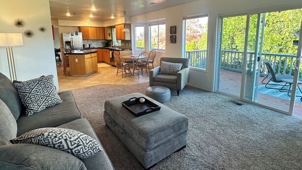 A spacious living room with a grey sofa, ottoman, and a dining area with chairs. A kitchen is visible in the background, and there is a large sliding glass door leading to an outdoor area. - Columbia View Retreat, Spacious Furnished Home in Argay Terrace,Portland Airport (Portland)