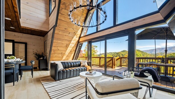 En rummelig loftsuite med stort vindue med udsigt over terrasse og bjerge. Den har en moderne sofa, spiseplads og en lysekrone.