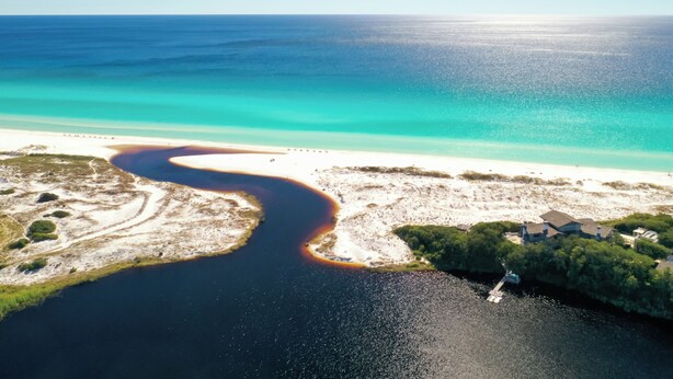Ett flygbildsbild av ett kustområde med en flod som leder till en strand, en brygga och ett hus med en stor trädgård.