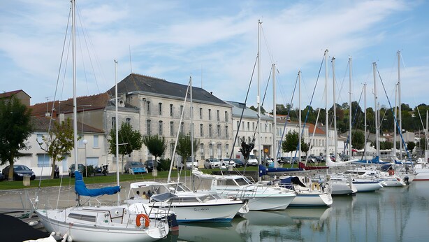 A marina with multiple boats docked, a large building in the background, and a calm water surface.