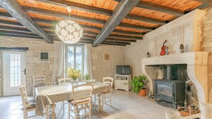 A rustic room with a stone wall, wooden beams, a fireplace, a dining table with chairs, and a television.