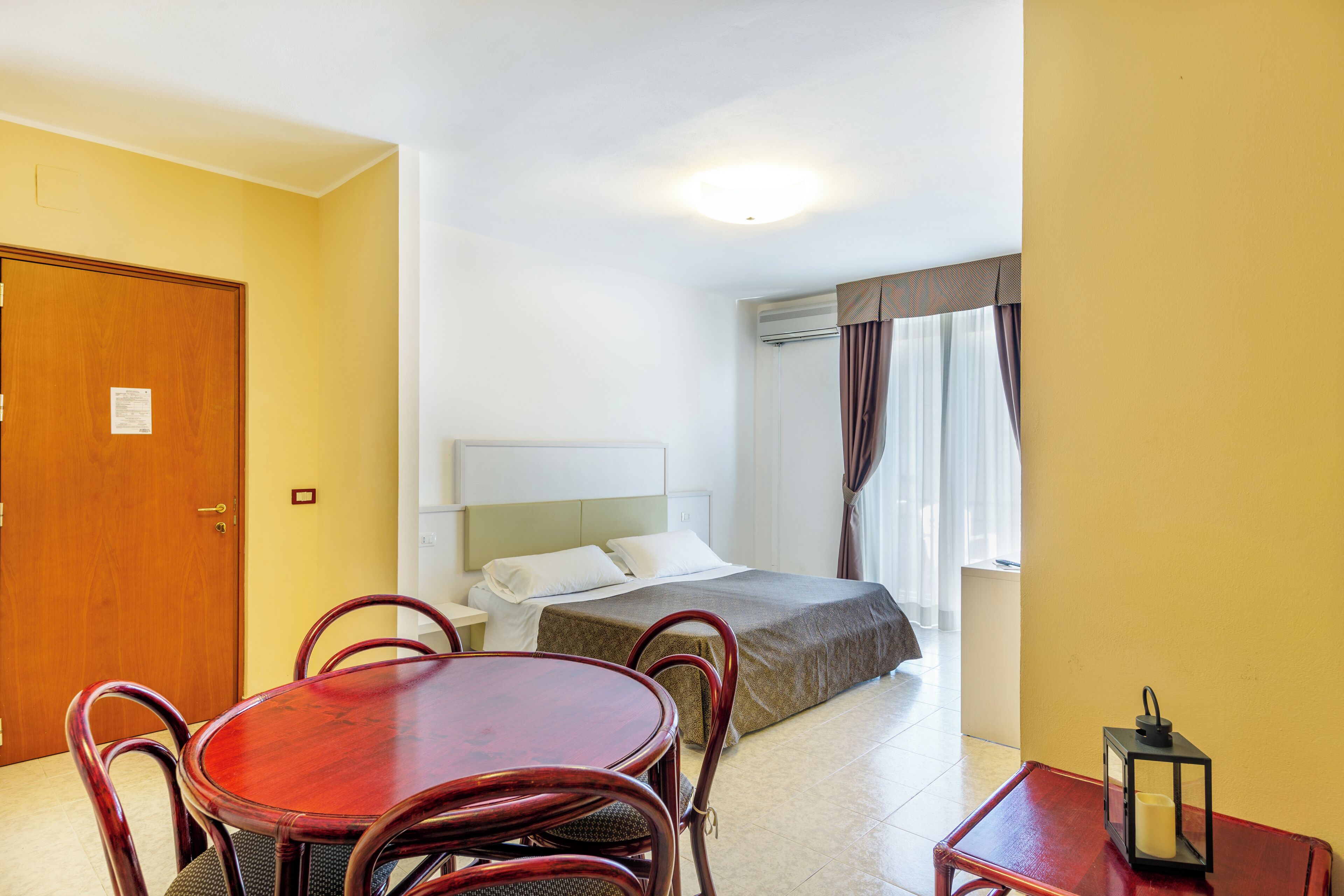 Habitación de hotel con una cama, una mesa de comedor de madera con sillas, una mesita auxiliar y una ventana con cortinas.
