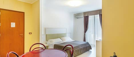 A hotel room with a bed, a wooden dining table with chairs, a small side table, and a window with curtains.