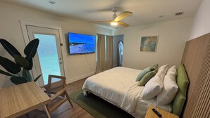 Una camera da letto con un letto, una scrivania, una sedia, una ventola a soffitto e una TV che mostra una spiaggia.