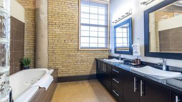 A modern bathroom with a brick wall, a large window, a bathtub, and a vanity with two sinks.