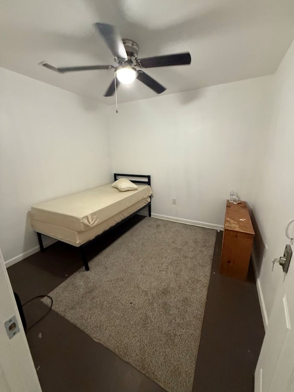 Una habitación pequeña con cama individual, ventilador de techo, mesita de madera y piso alfombrado.