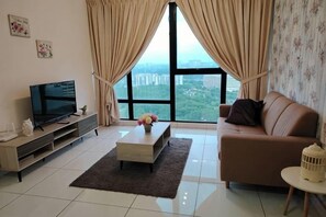A living room with a sofa, a coffee table, a TV on a stand, and a view through large windows. - City View Southkey 2BR 2FREE 18F By Natol (Plentong)