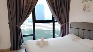 A hotel room with a bed, curtains, a window with a view of buildings, and a headboard. - City View Southkey 2BR 2FREE 18F By Natol (Plentong)
