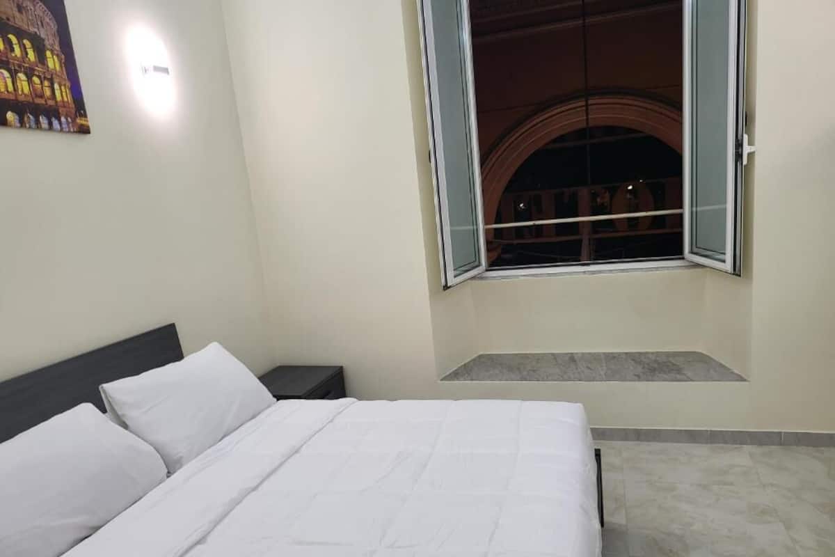 A bedroom with a bed, a nightstand, a window with an open view, and a small seating area.