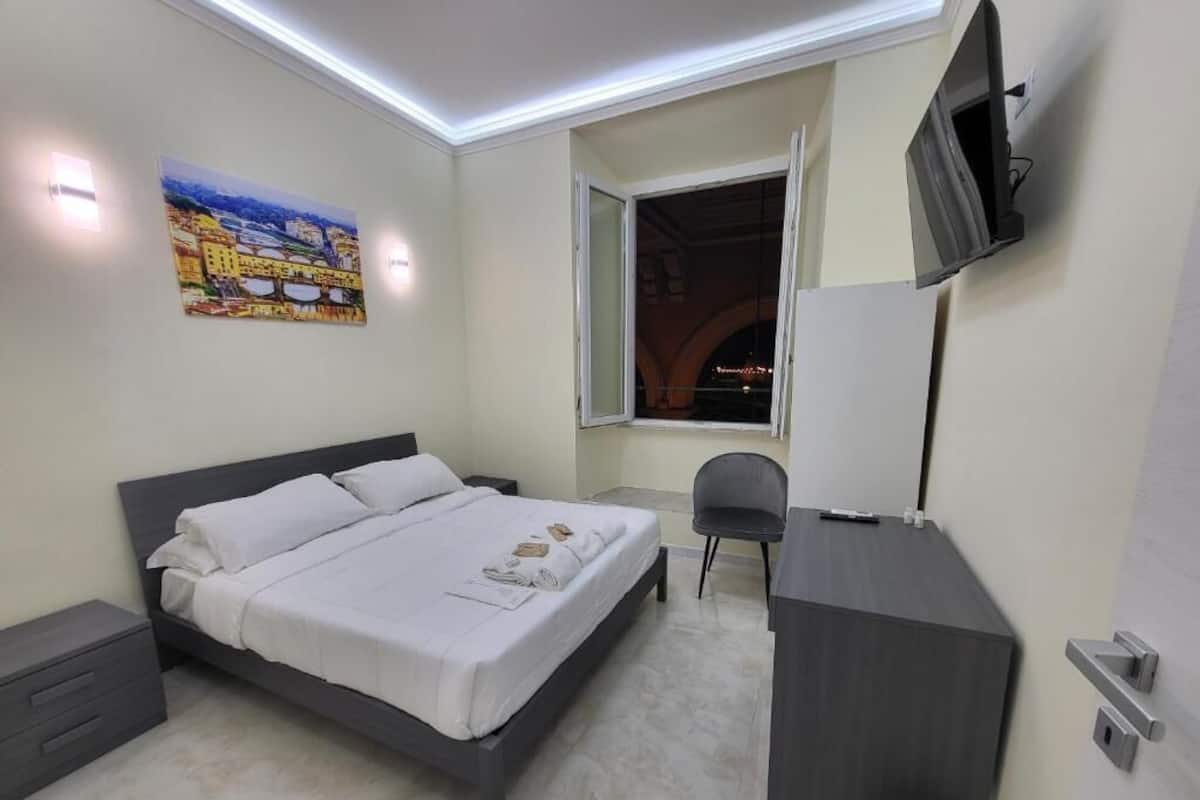 A bedroom with a bed, a desk, a chair, a TV, and a window with an open view.