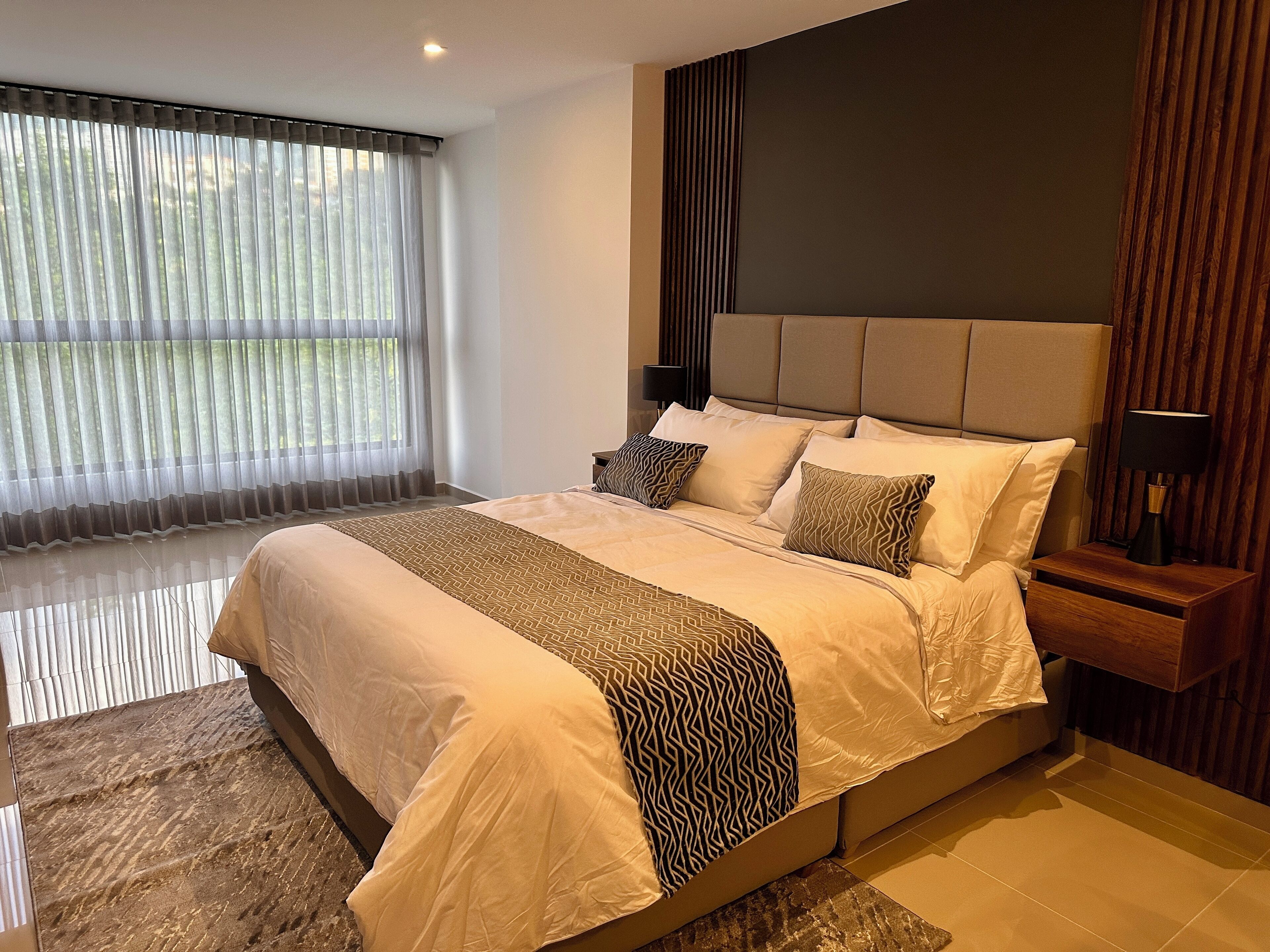 Een moderne slaapkamer met een groot bed, een nachtkastje met een lamp, en uitzicht op groen door dunne gordijnen.