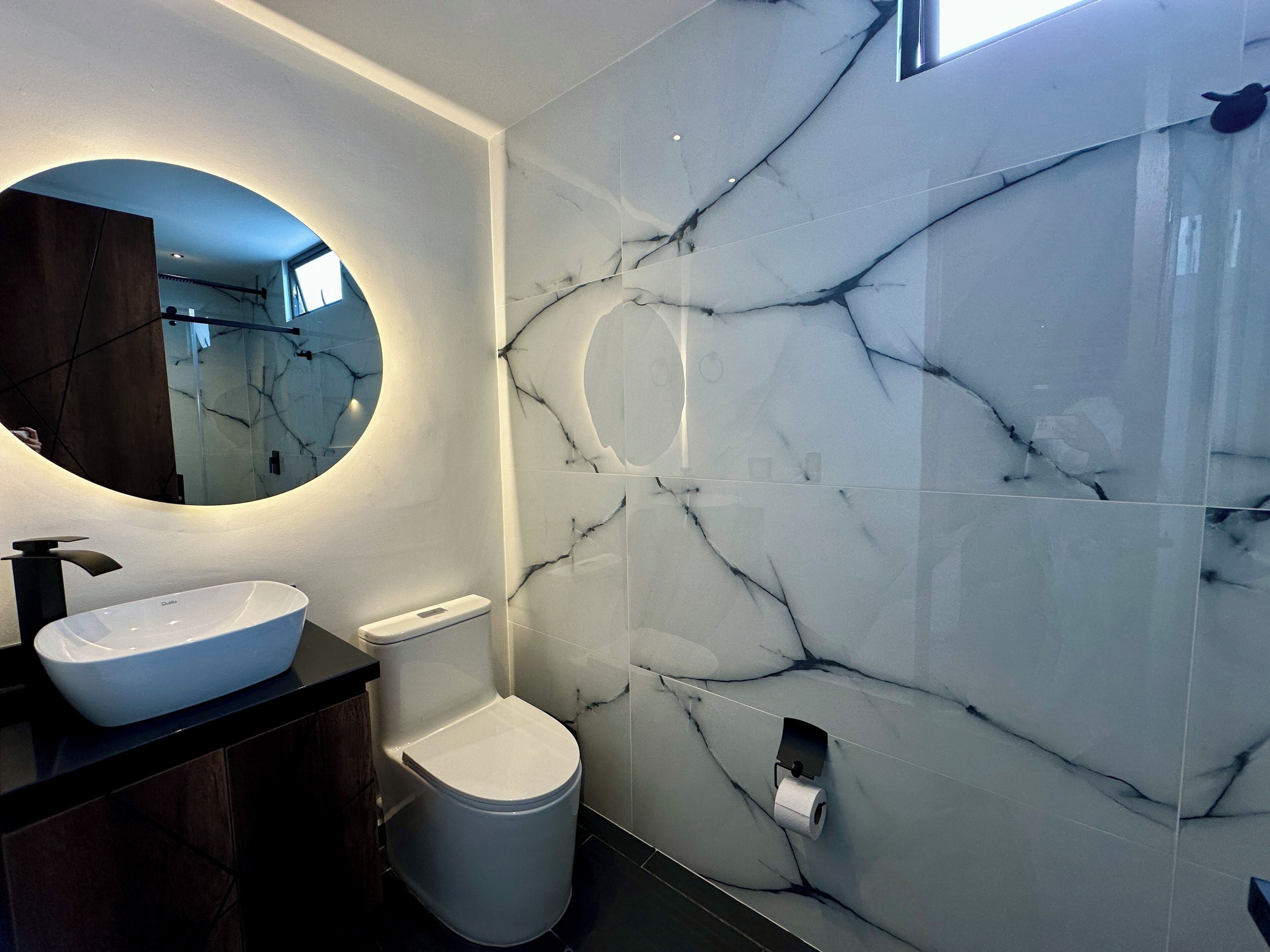 A modern bathroom with a white marble sink, a toilet, and a mirror with a circular frame.