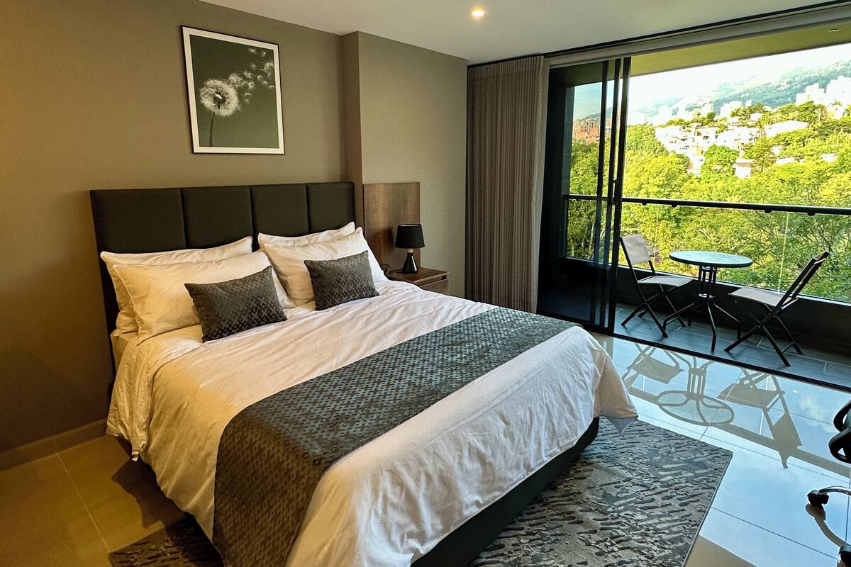 A large bed with a headboard, a bedside table with a lamp, a framed picture on the wall, and a balcony with a table and chairs.