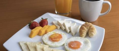 A breakfast plate with eggs, fruit, and pastries on a white plate, a glass of orange juice, and a cup of coffee on a wooden table.