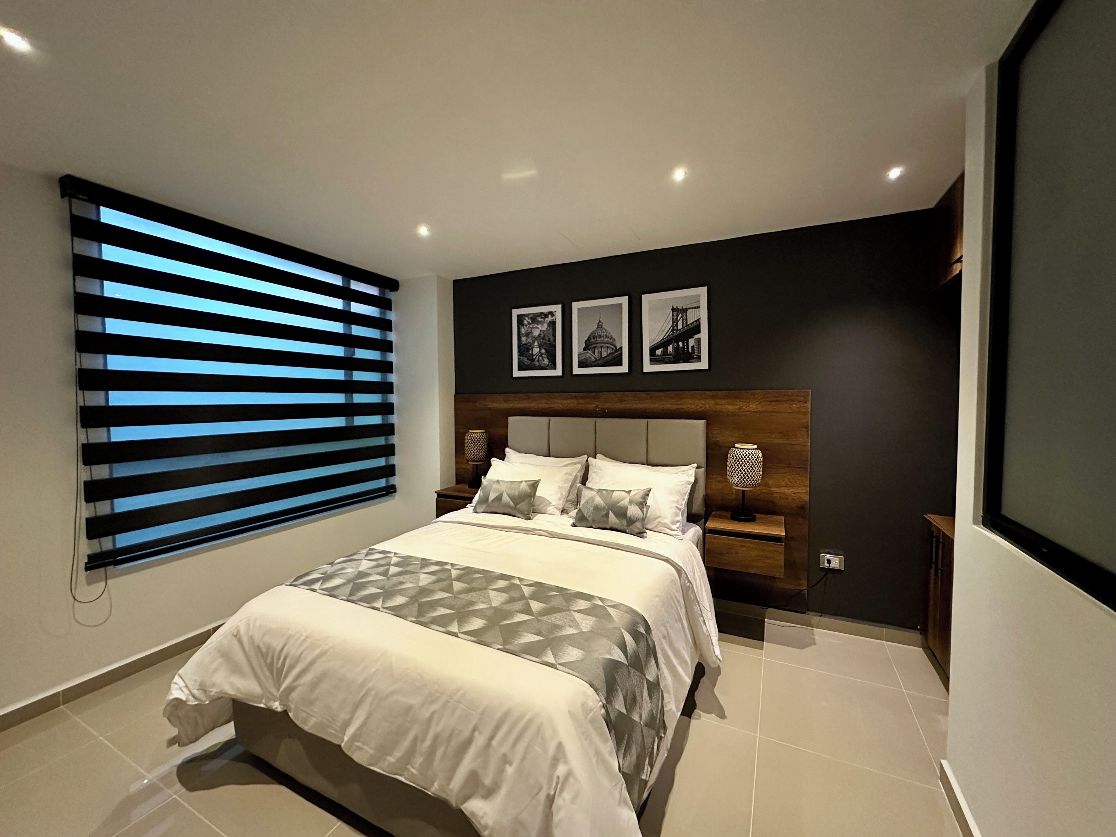 Quarto moderno com uma cama grande, cabeceira com três quadros, mesa de cabeceira com luminária e janela com persianas.