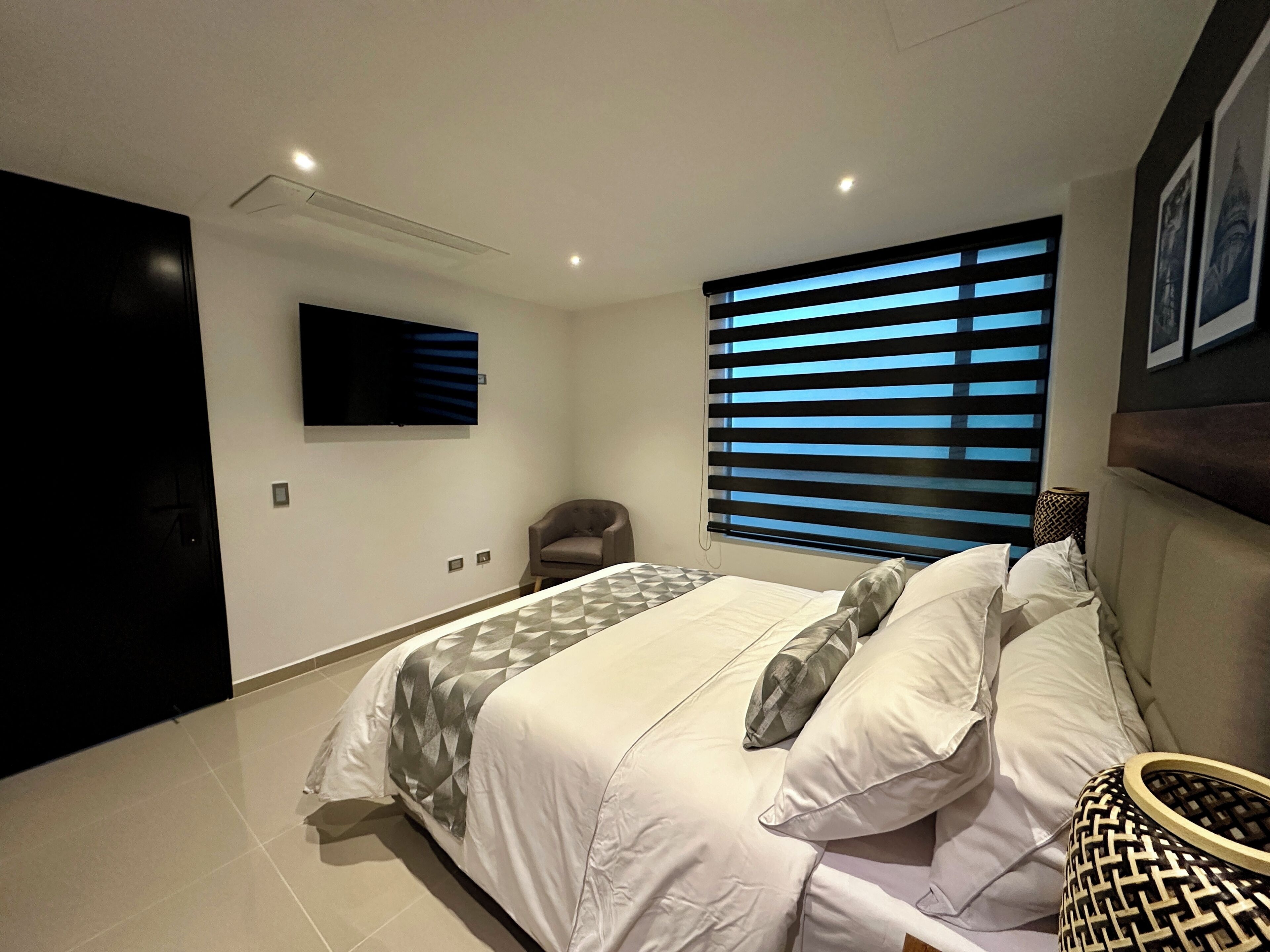 Een moderne slaapkamer met een groot bed, een flatscreen televisie aan de muur en een raam met horizontale zonwering.