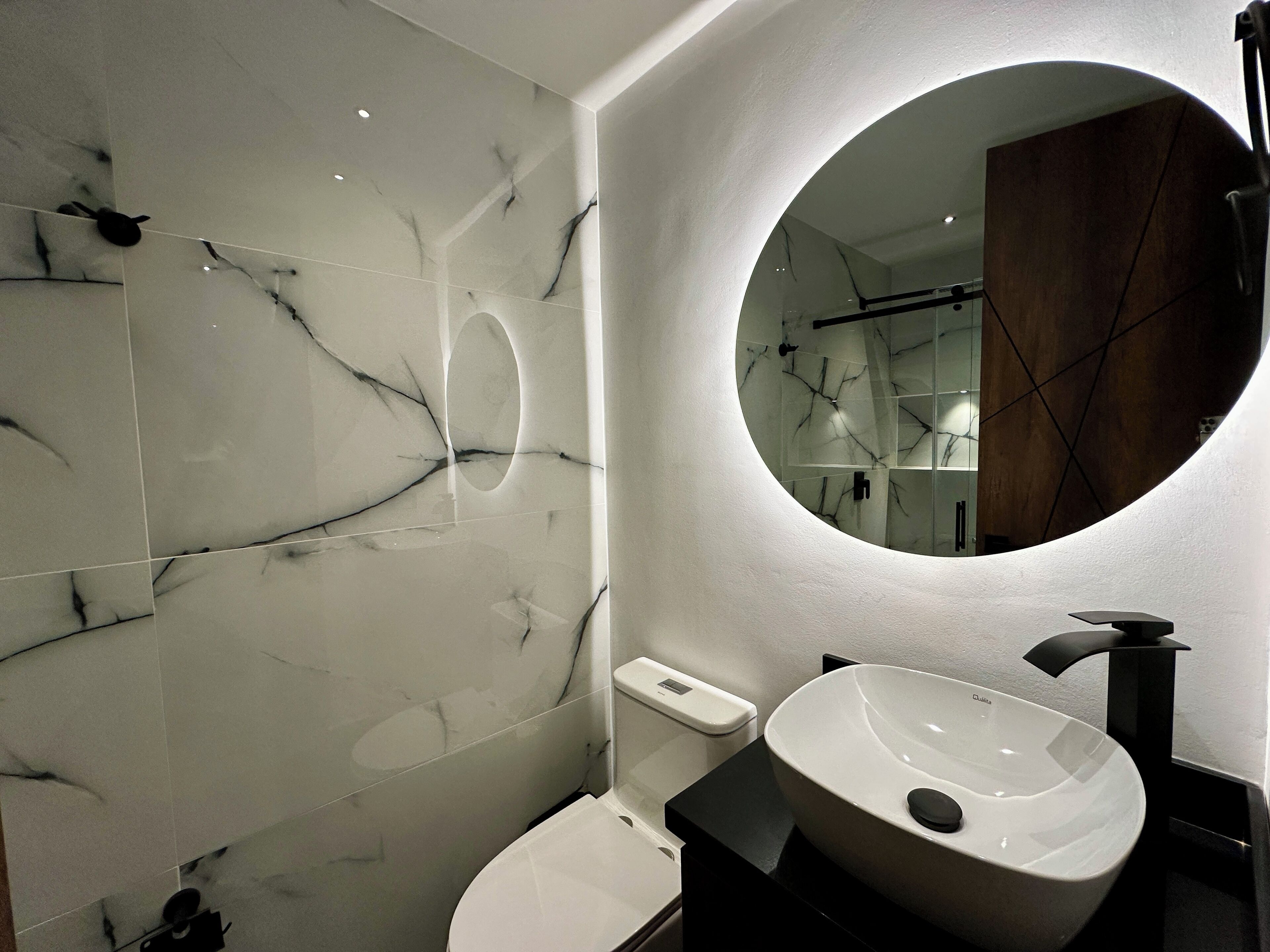 A modern bathroom with a white marble wall, a round mirror, a white sink, and a black countertop.