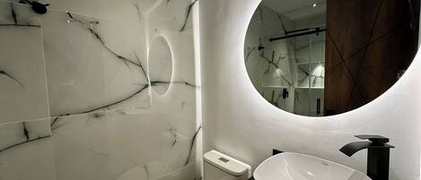 Un baño moderno con pared de mármol blanco, un espejo redondo, un lavabo blanco y encimera negra.