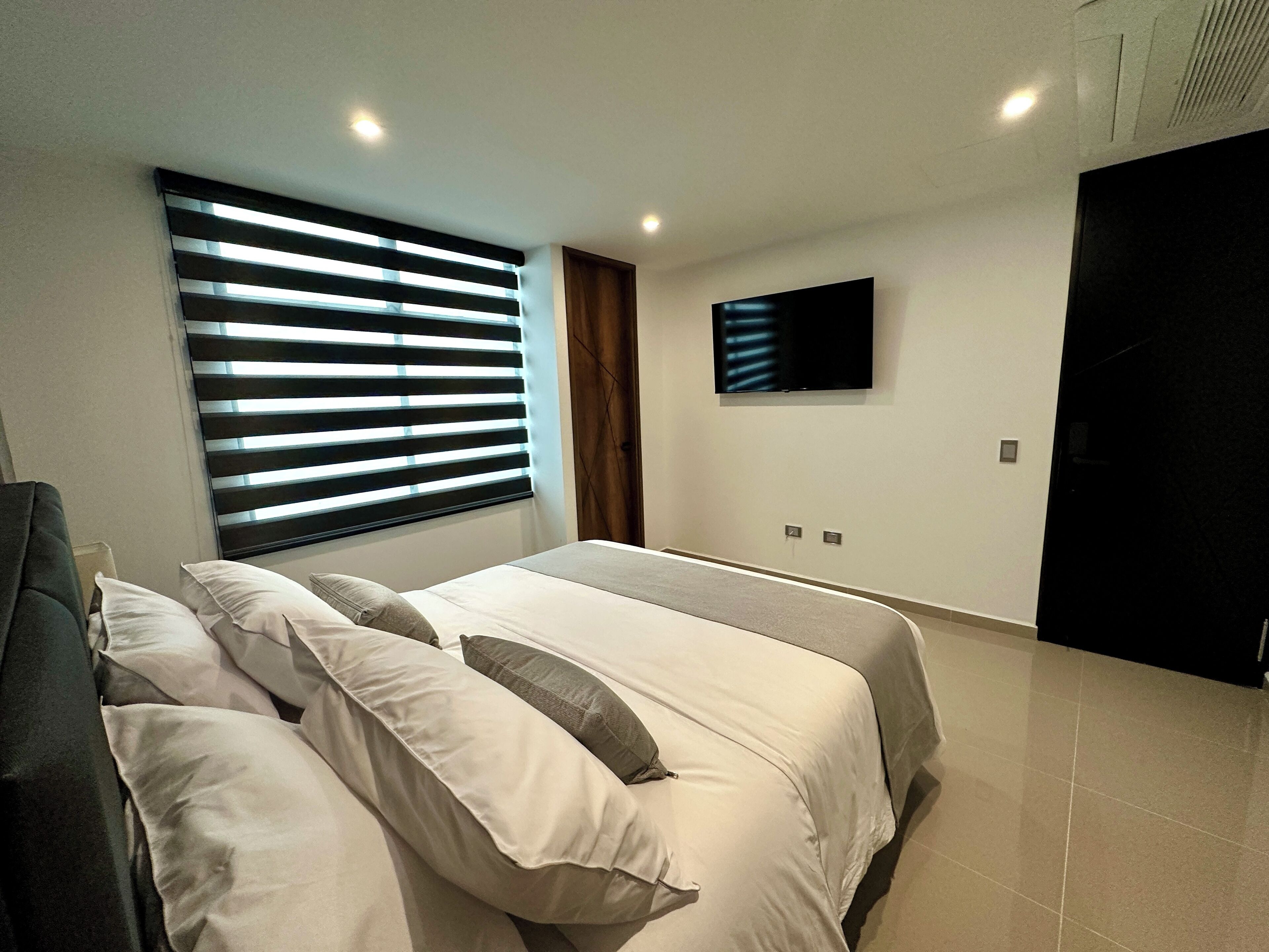 Une chambre à coucher moderne avec un lit, un téléviseur fixé au mur et une fenêtre avec des stores.