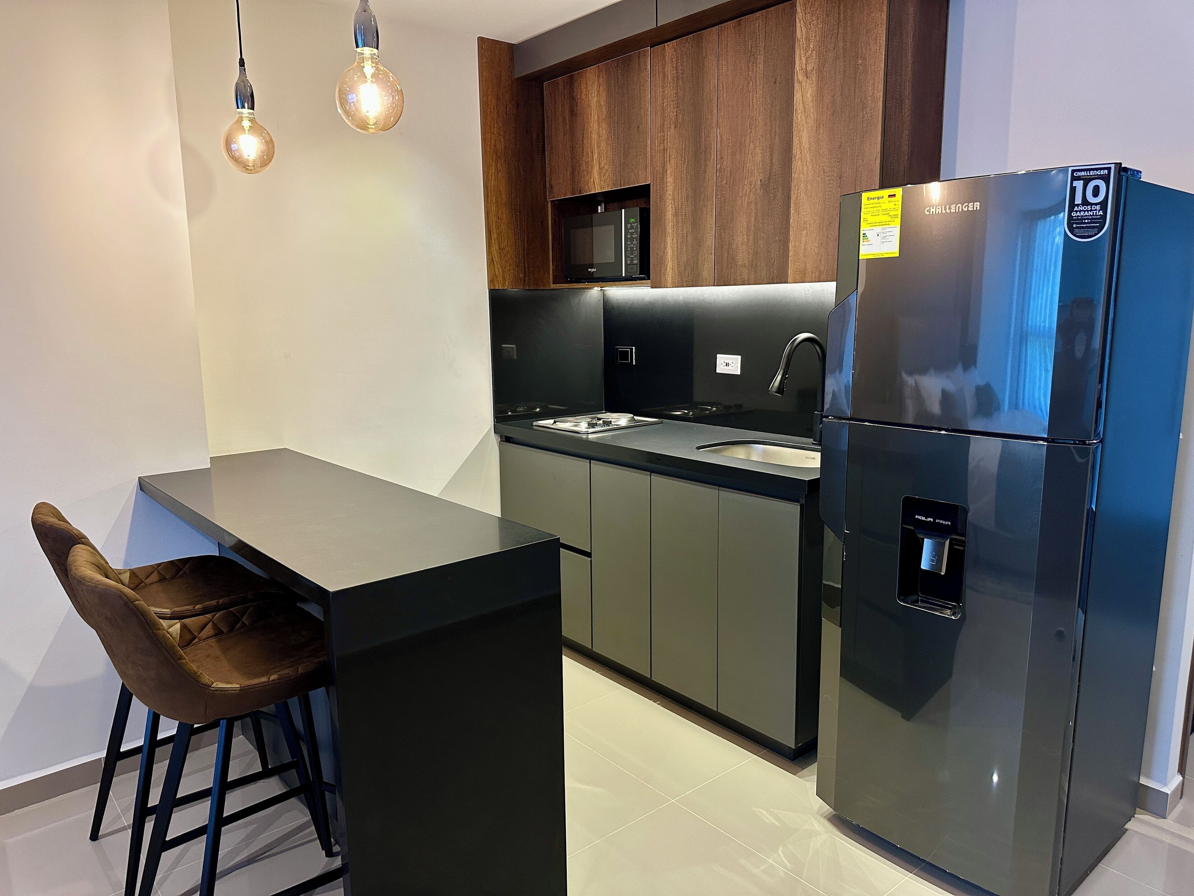 Una cocina moderna con un refrigerador negro, un microondas y un lavamanos. Hay dos lámparas colgantes y un taburete de bar.