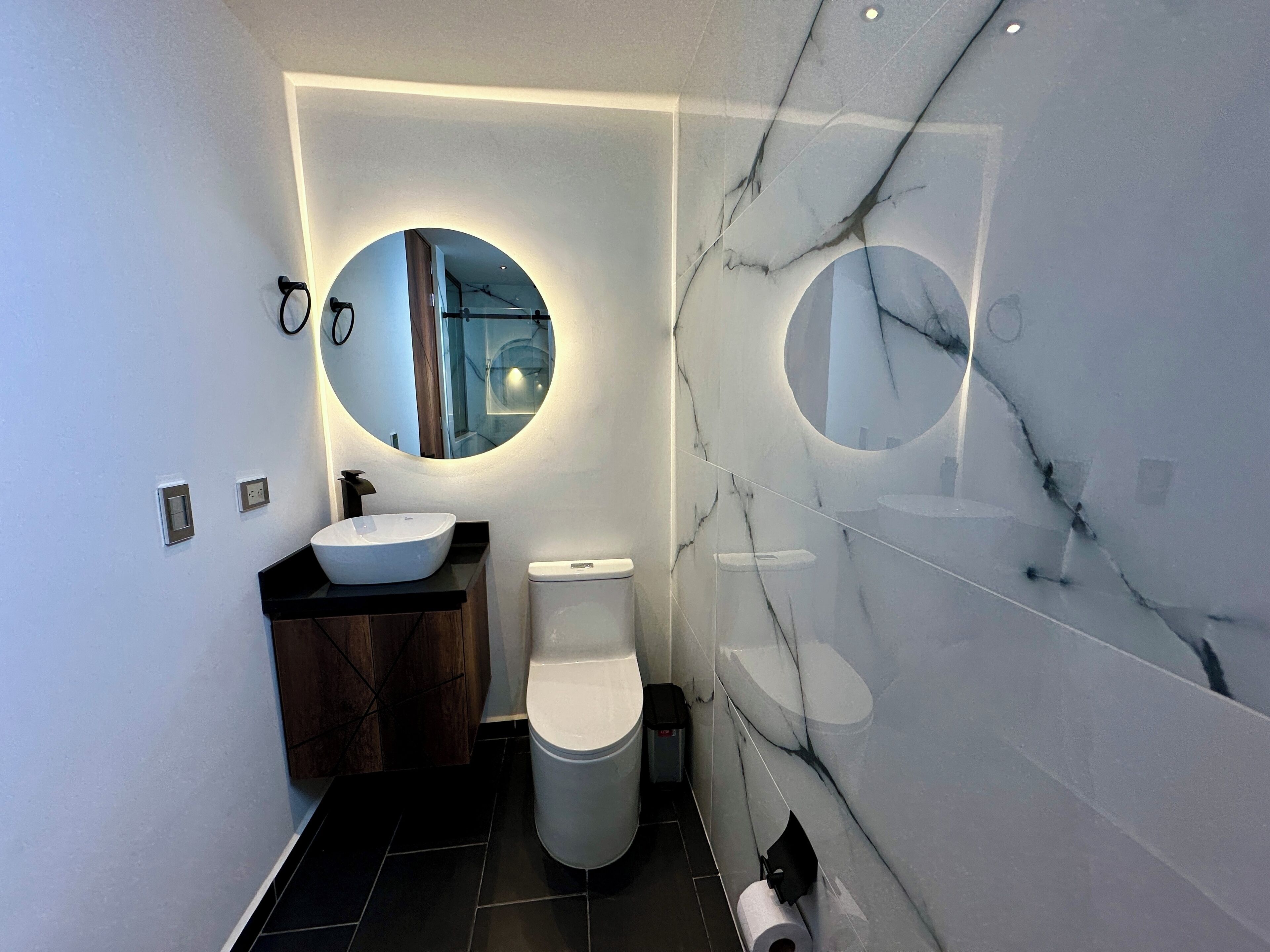 A modern bathroom with a round mirror, a vessel sink, a toilet, and a marble wall.