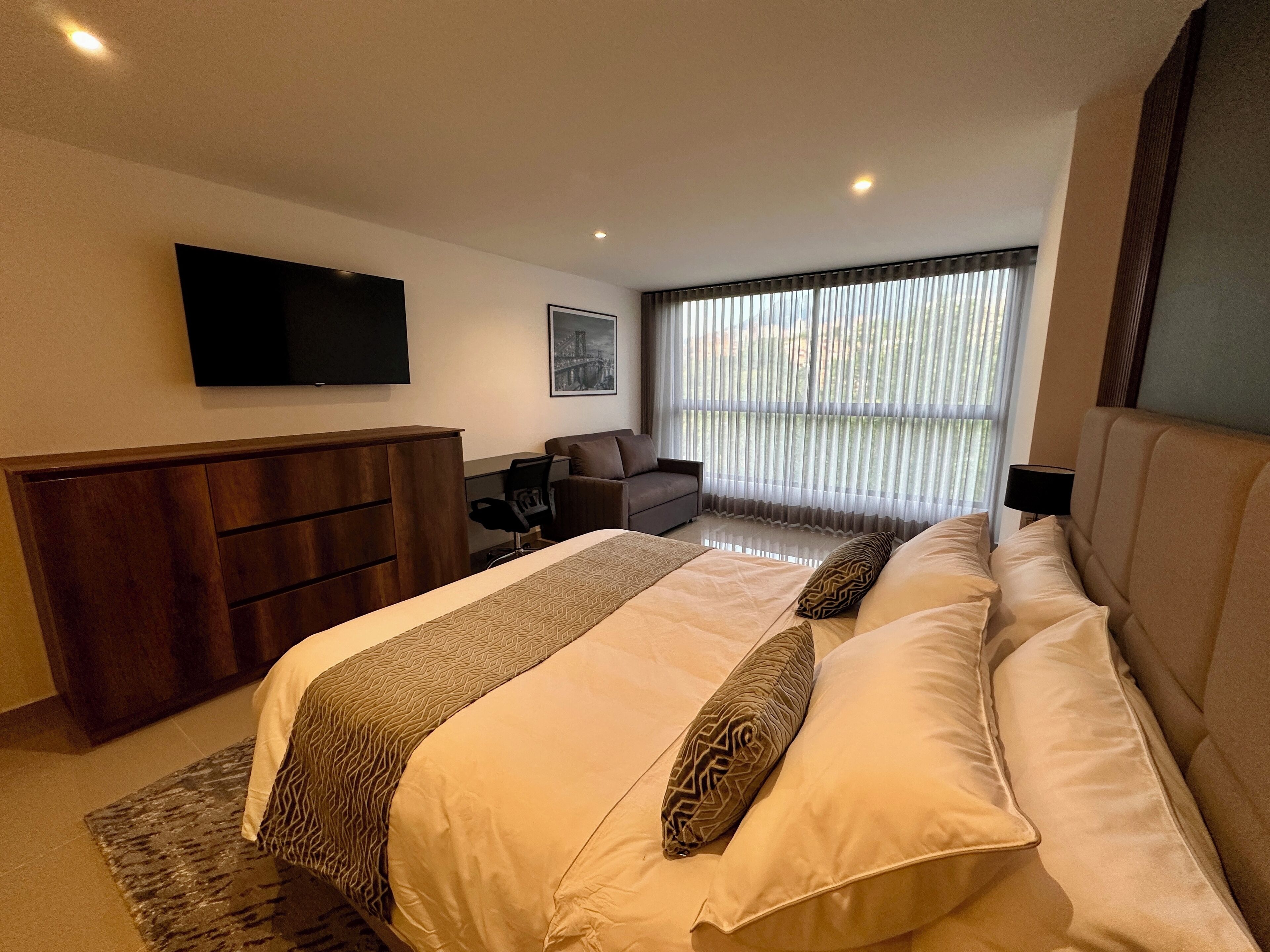 A modern hotel room with a large bed, a desk, a sofa, and a TV mounted on the wall.