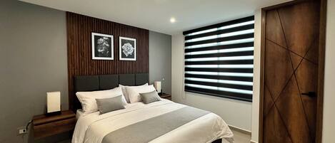 A modern bedroom with a bed, bedside tables, a nightstand, a wall-mounted light, a window with blinds, and a door.