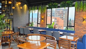 Um café moderno com mesas e cadeiras de madeira, assentos acolchoados em azul, plantas penduradas e grandes janelas com vista para a cidade.