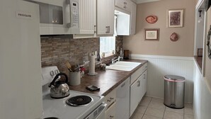 Una cocina con alacenas blancas, microondas, estufa y lavamanos. Hay elementos decorativos en la encimera y un bote de basura en la esquina.