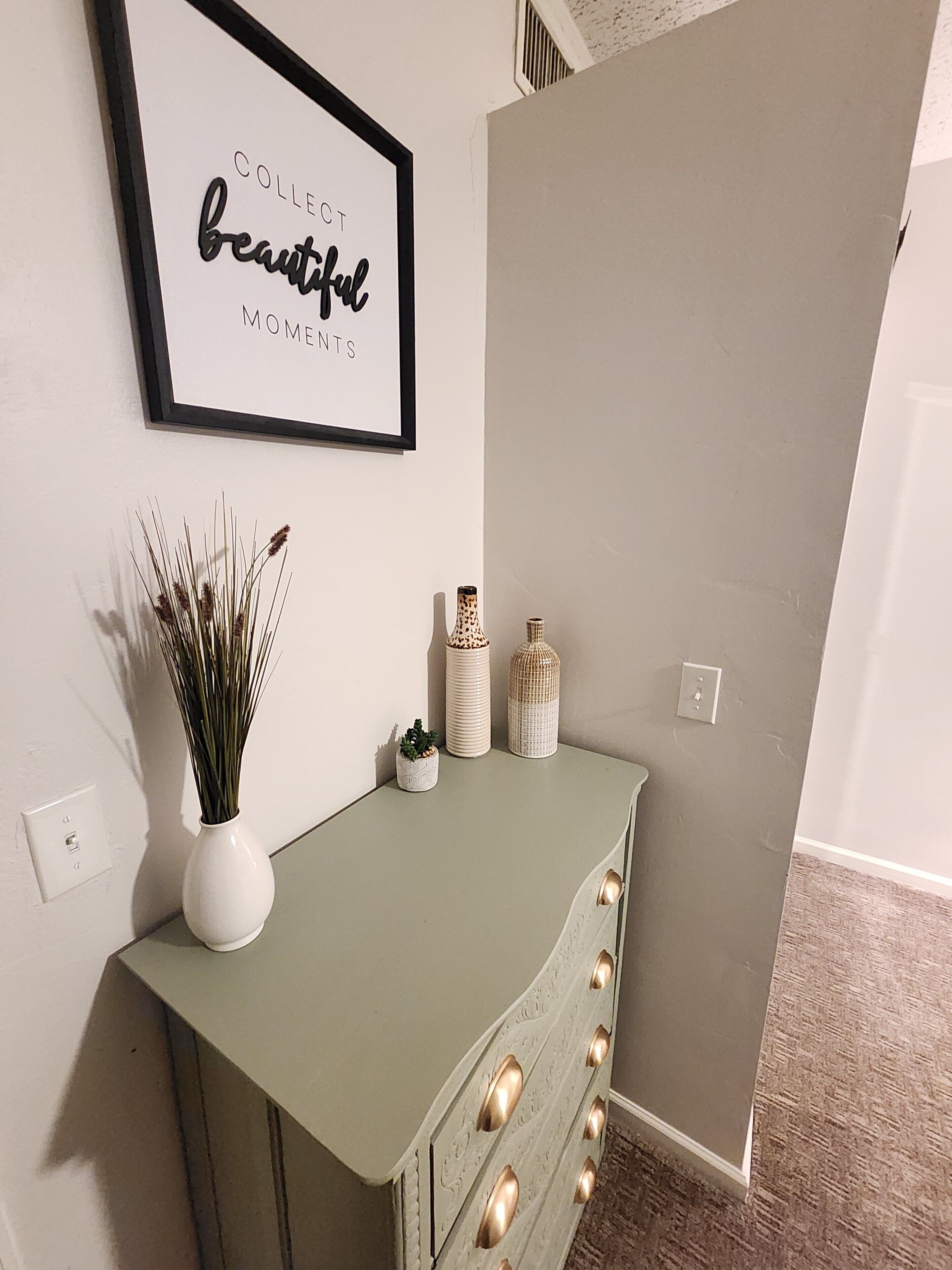 Ein Zimmer mit einem grünen Schminktisch, einem gerahmten Schild an der Wand und einer kleinen Pflanze auf dem Schminktisch.