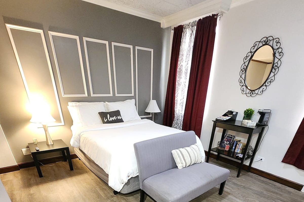 A bedroom with a bed, bedside tables, a bench, a nightstand, a mirror, and a small table with a plant and books.