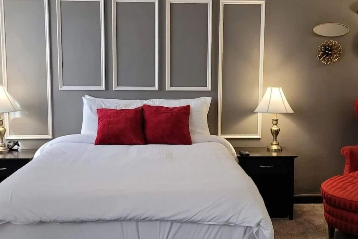 A neatly made bed with white and red pillows, a black nightstand with a lamp, and a red chair.