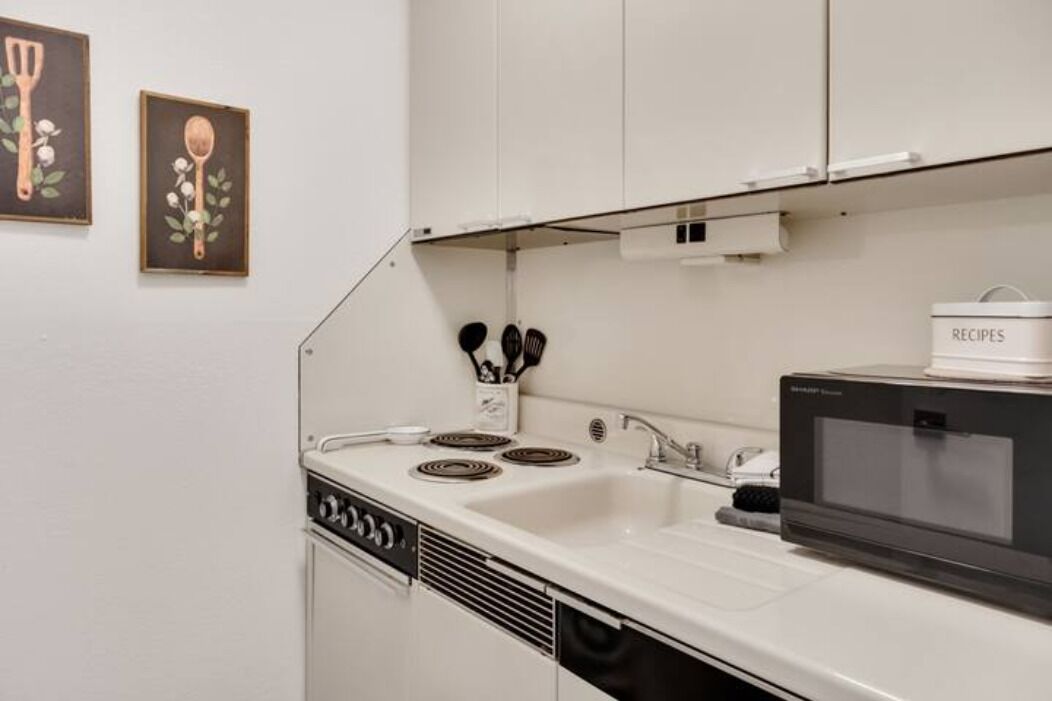 Una cocina compacta con armarios blancos, estufa, fregadero y microondas. Hay dos cuadros en la pared.