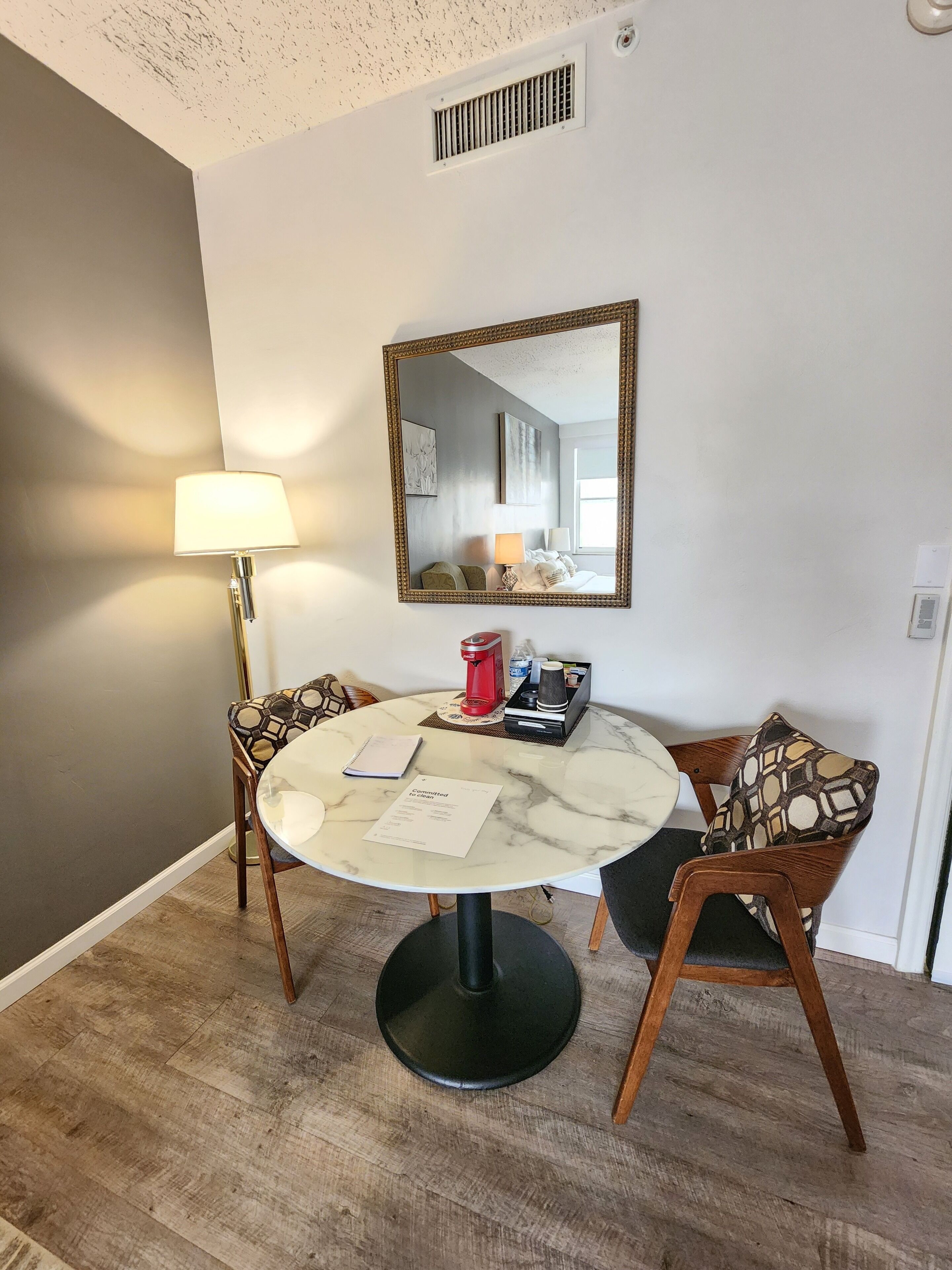 A dining area with a round table, chairs, a lamp, a mirror, and a coffee maker.