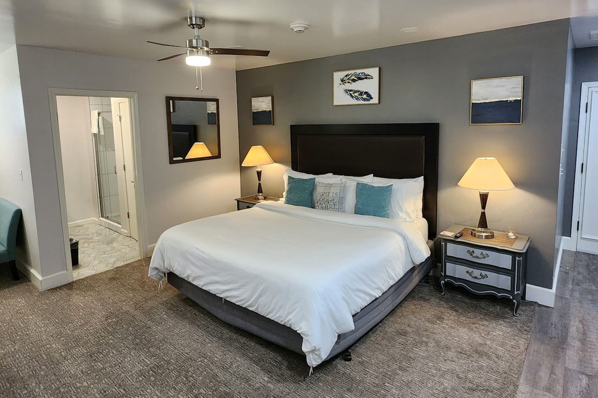 A bedroom with a large bed, two bedside tables with lamps, a ceiling fan, and a door leading to a bathroom.