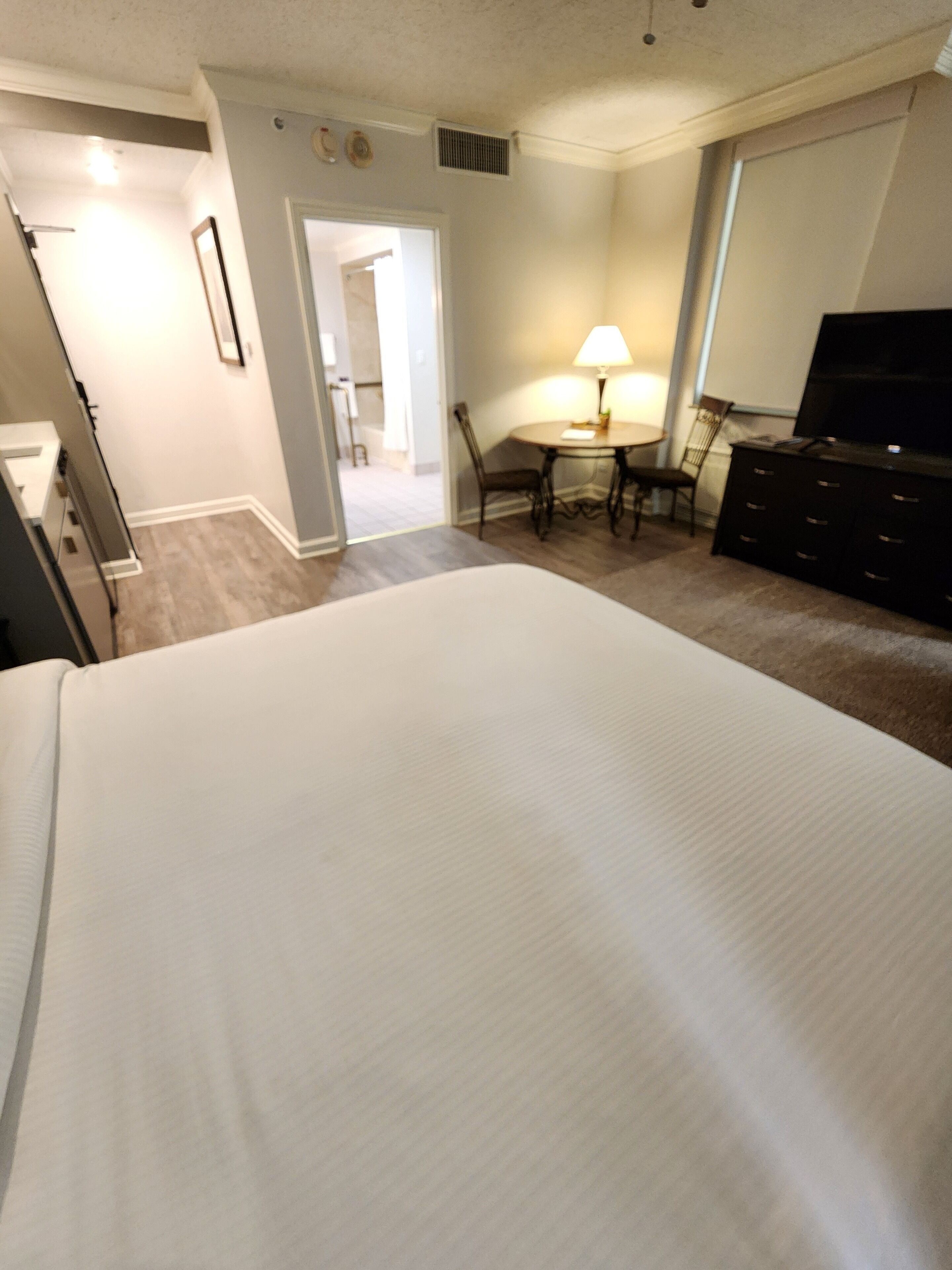 A hotel room with a large bed, a desk with a lamp, a dresser, and a TV.