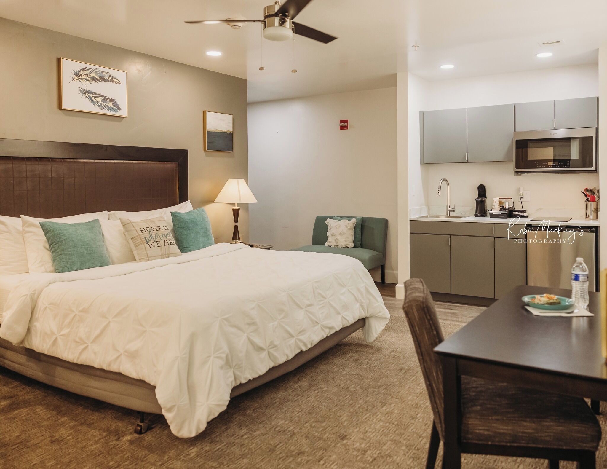 A hotel room with a bed, a desk, a chair, a lamp, a green armchair, a kitchen area with a microwave, and a ceiling fan.
