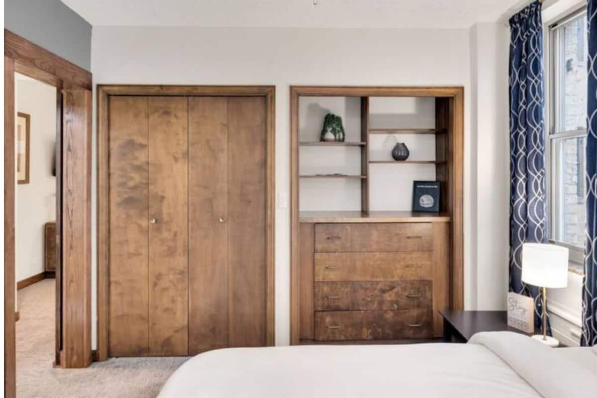 A bedroom with a wooden wardrobe, a built-in shelf unit, a bed, and a nightstand with a lamp.