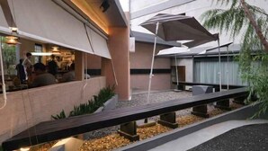 Zona salotto all'aperto con tavoli e ombrelloni, circondata da un edificio moderno con tetto spiovente e vialetto ghiaioso.
