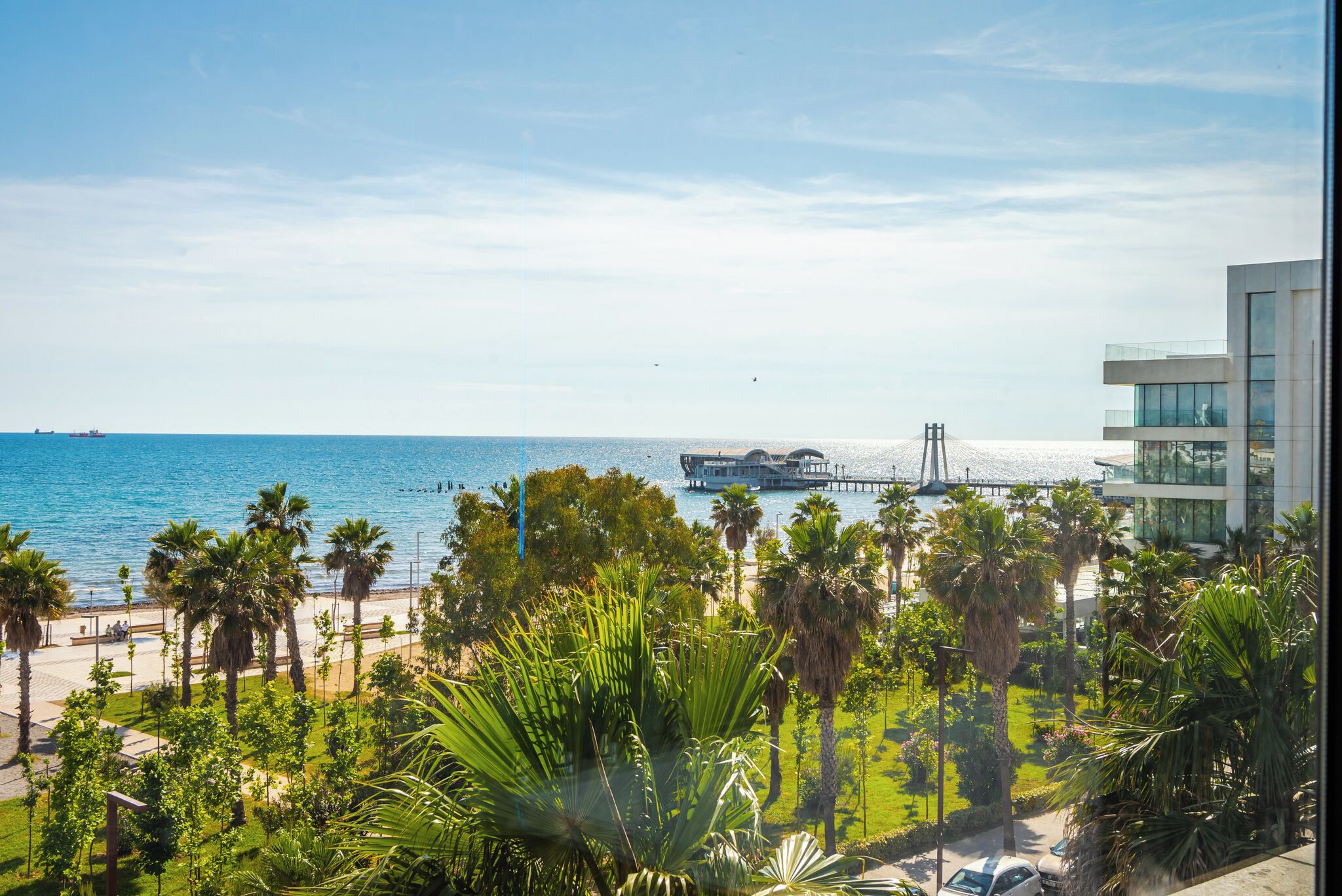 A coastal hotel with a view of the sea, palm trees, and a modern building with glass windows.
