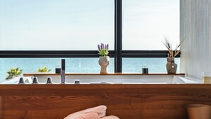 Un baño moderno con tocador de madera, un ventanal con vistas al mar y una pequeña planta en maceta sobre el alféizar.