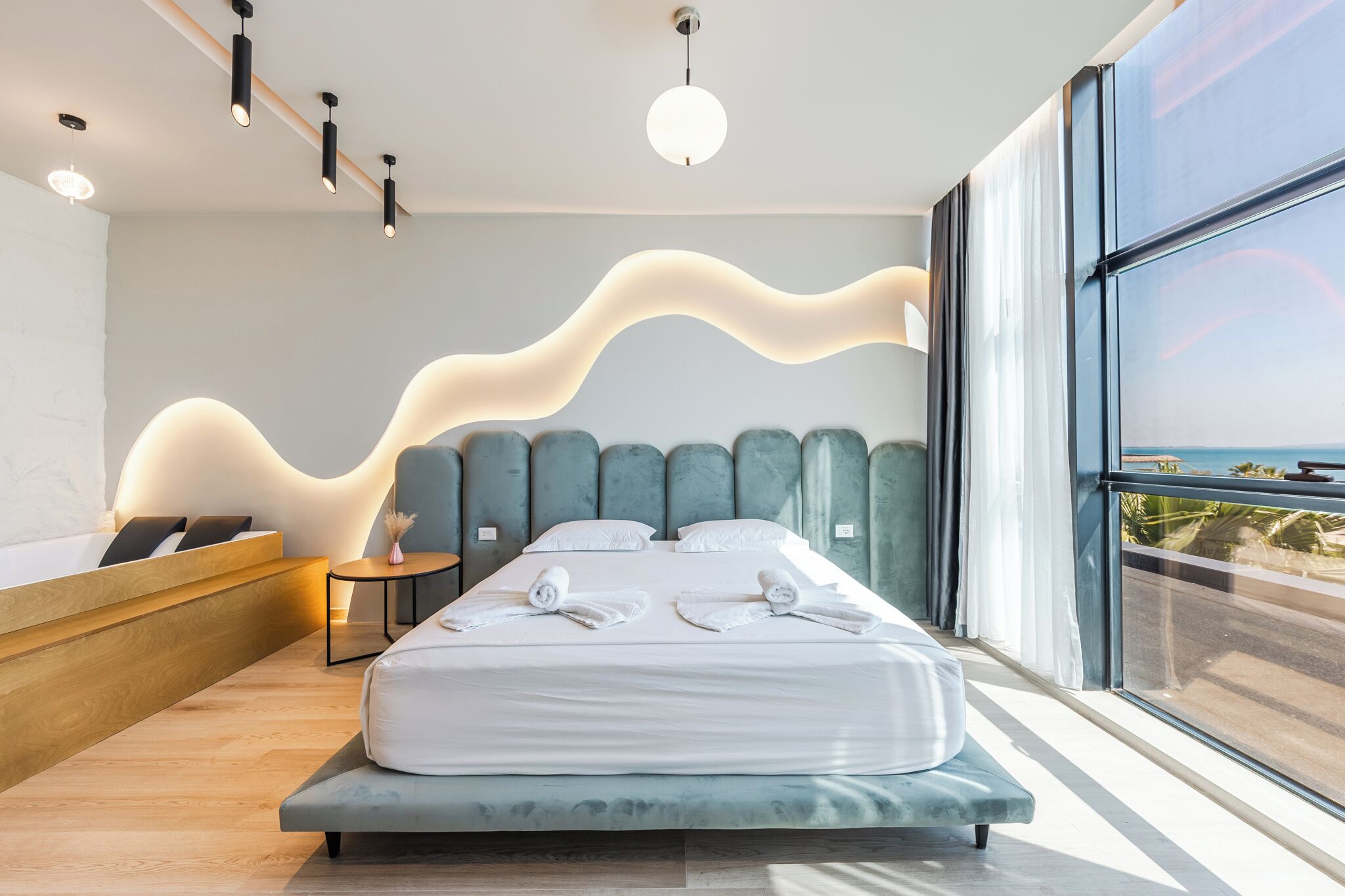 Habitación de hotel moderna con una cama grande, un banco de madera, una mesita auxiliar y vista al océano a través de grandes ventanales.