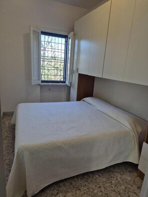 Une petite chambre avec un lit simple, une fenêtre avec des barreaux et une armoire.
