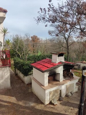 Un espace barbecue extérieur en béton, doté d’un toit rouge, entouré de verdure et d’une clôture.