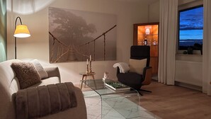 Un soggiorno con divano, poltrona e tavolino in vetro. Una tela raffigura un albero sulla parete, una lampada e vista mare attraverso una finestra.